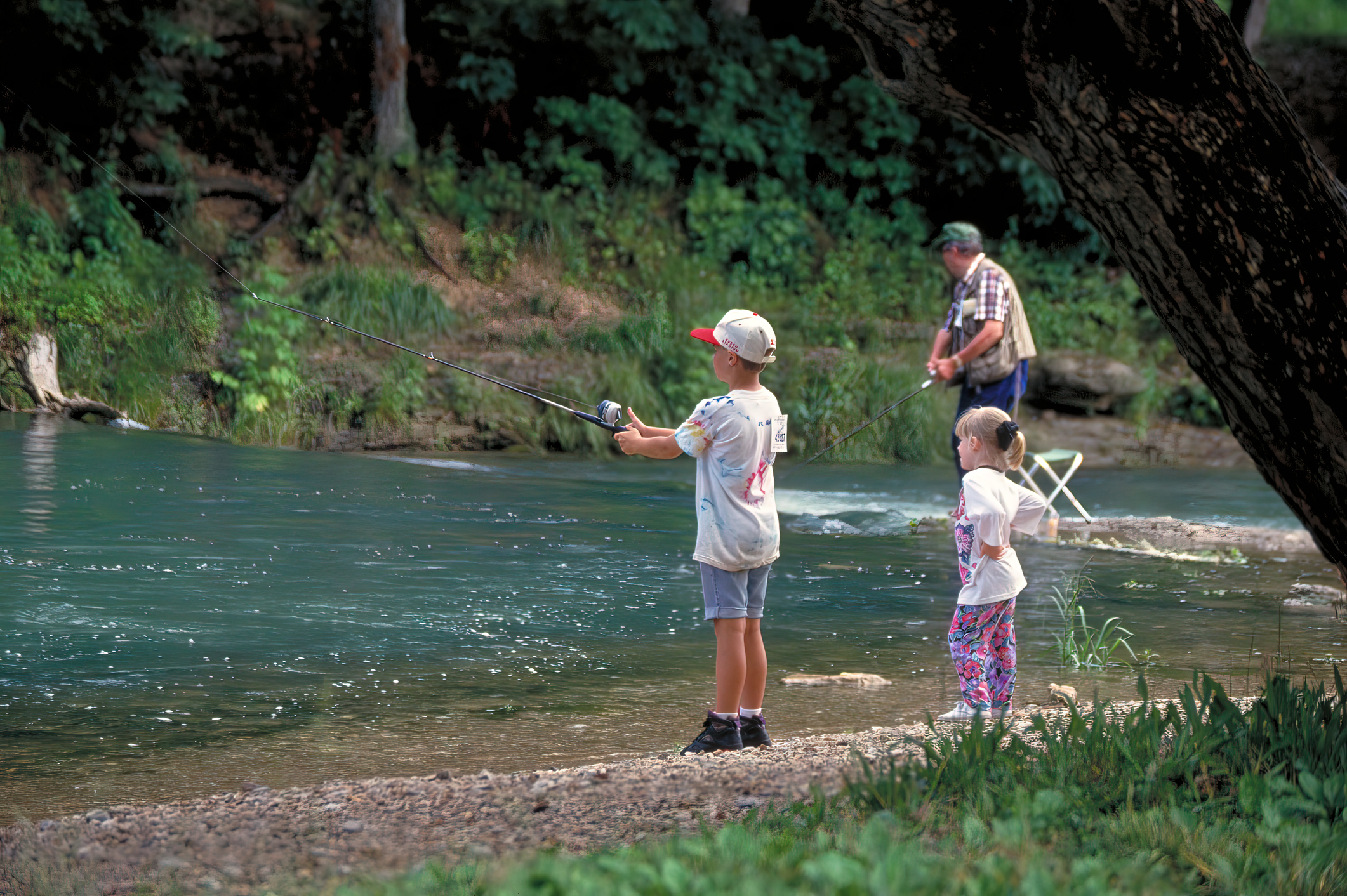 A boy and girl fishing by a riverbank under a large tree, with an adult fishing in the background.