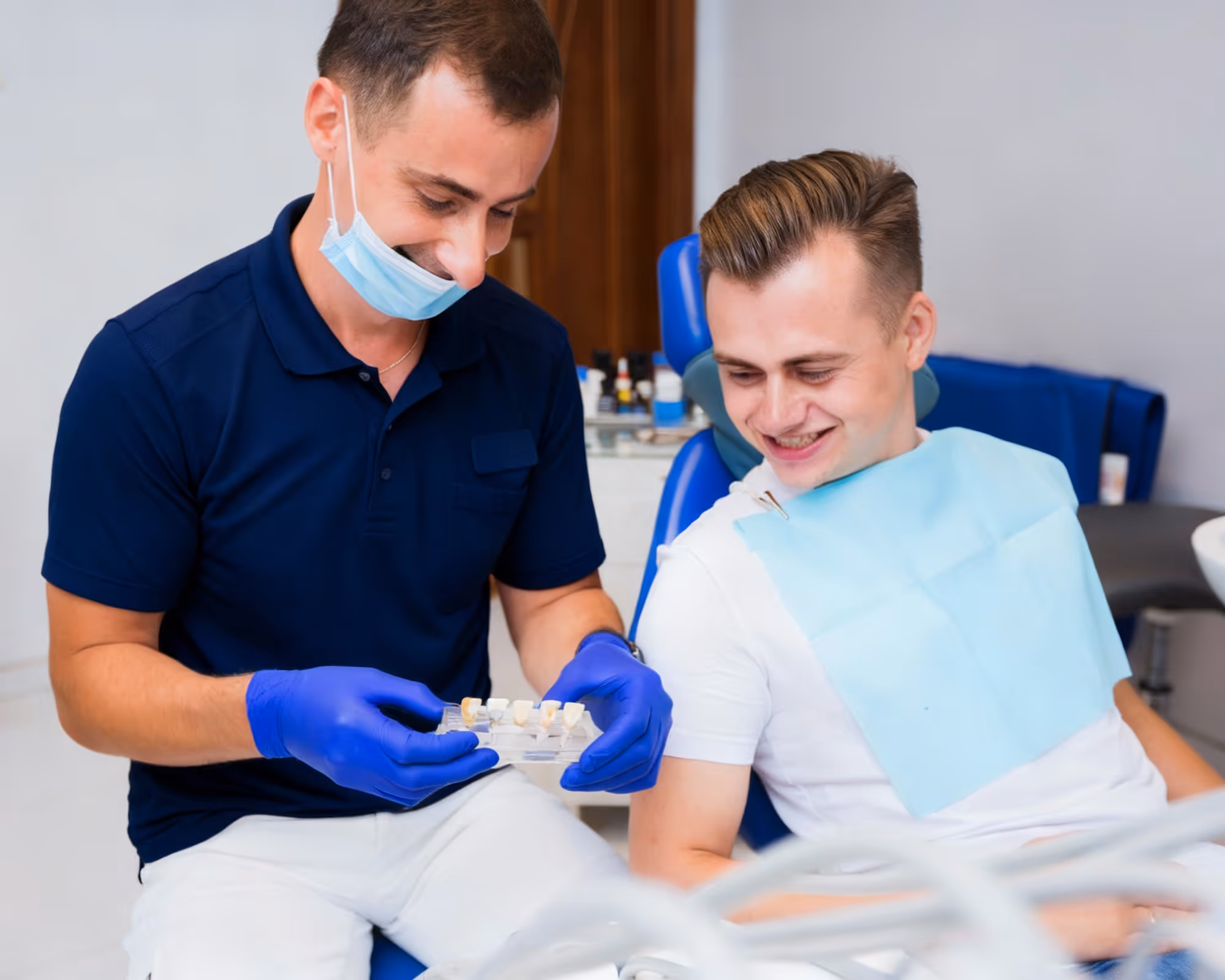 Dentist wearing blue gloves and mask shows dental crowns to a smiling male patient seated in a dental chair.