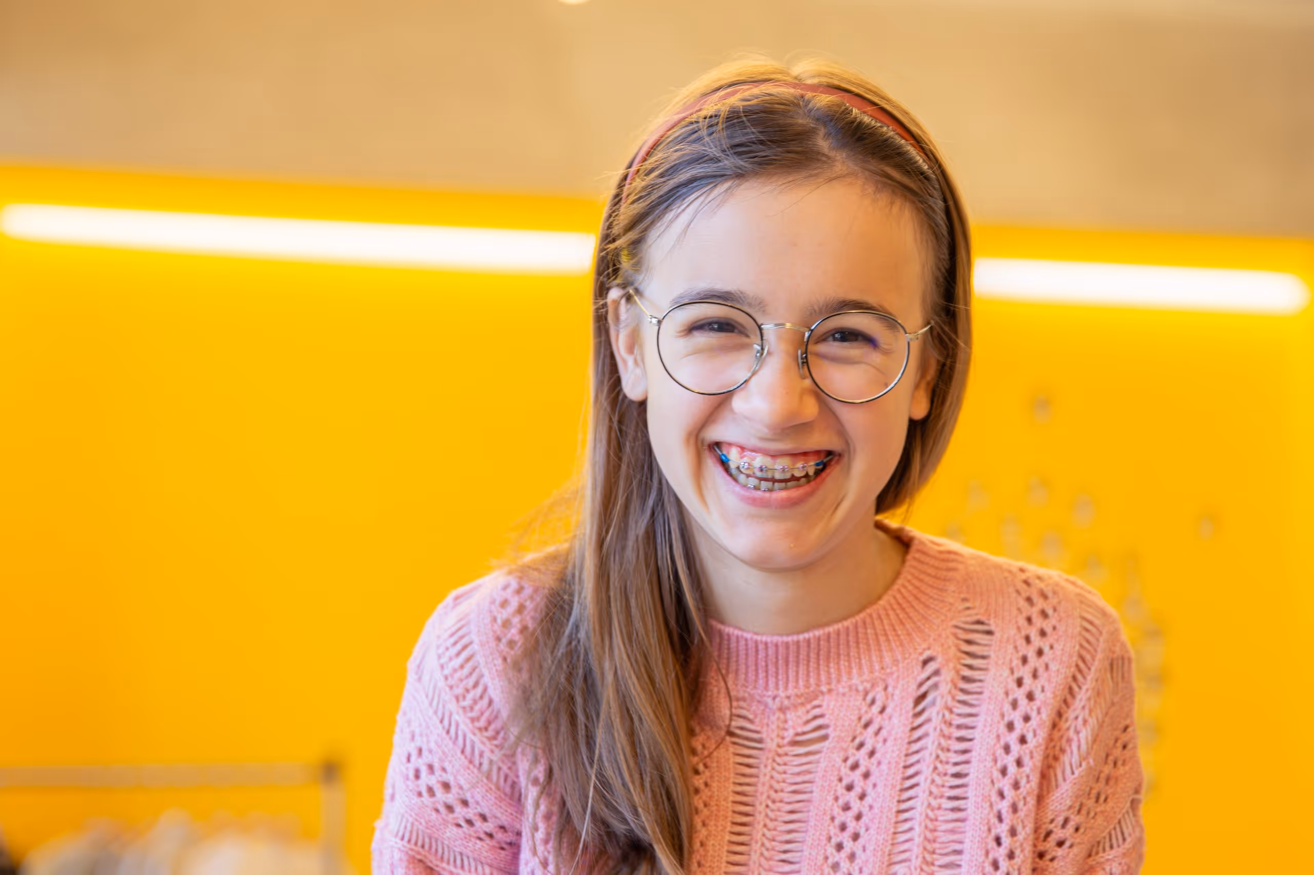 Smiling young woman with braces wearing glasses and a pink knitted sweater against a bright yellow background.