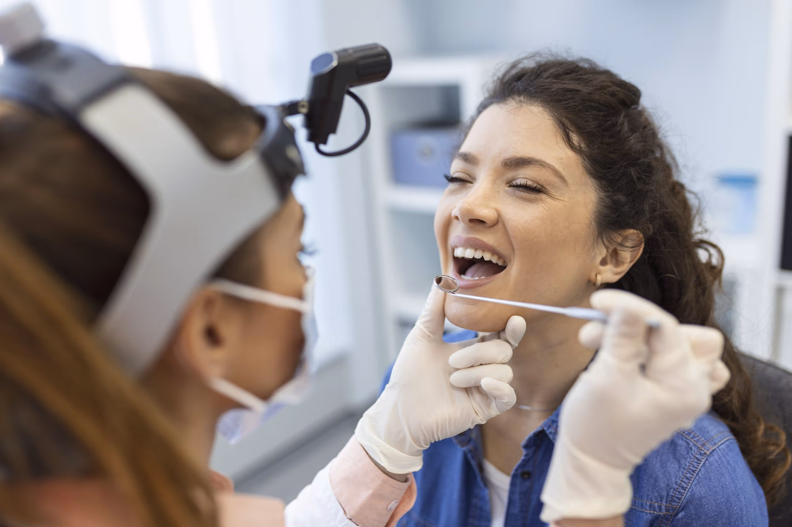 Dentist wearing gloves and a head mirror examining a smiling woman's teeth with a dental mirror.