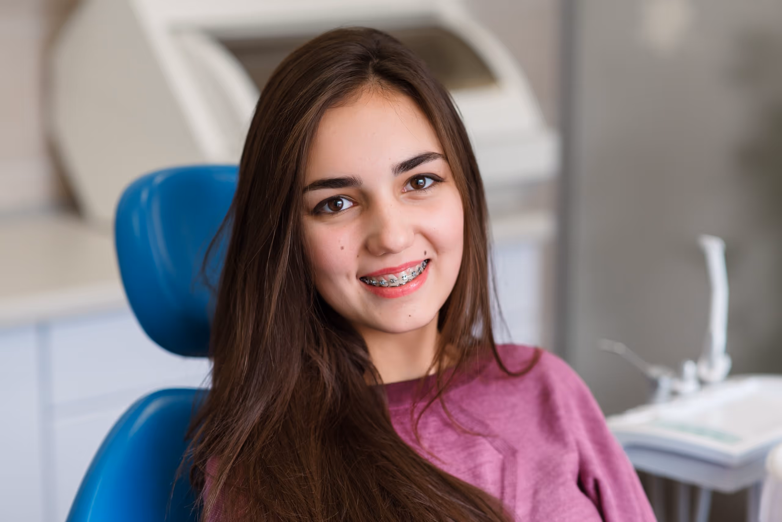 Smiling young woman with long brown hair and braces sitting in a dental chair.