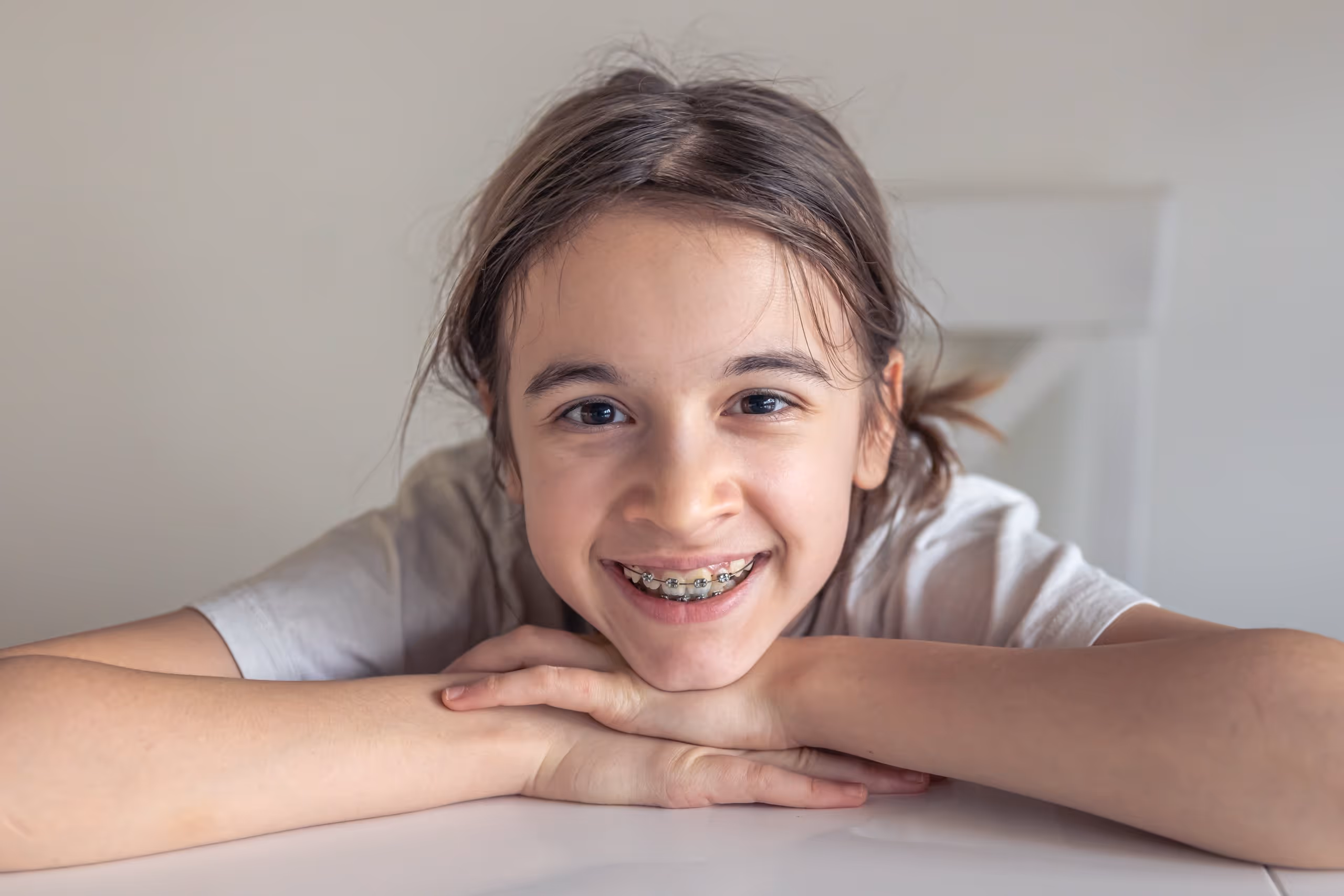 Smiling girl with braces resting her chin on folded hands on a white surface.