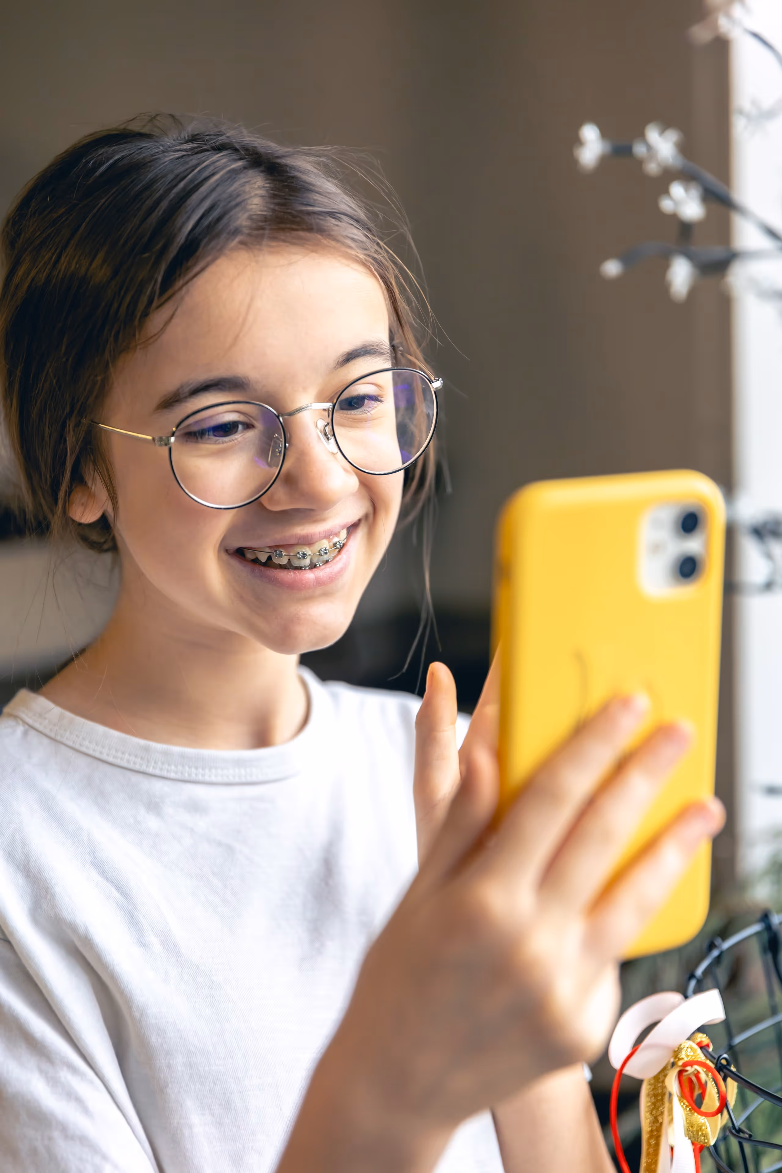 Smiling girl with glasses and braces looking at a yellow smartphone indoors.