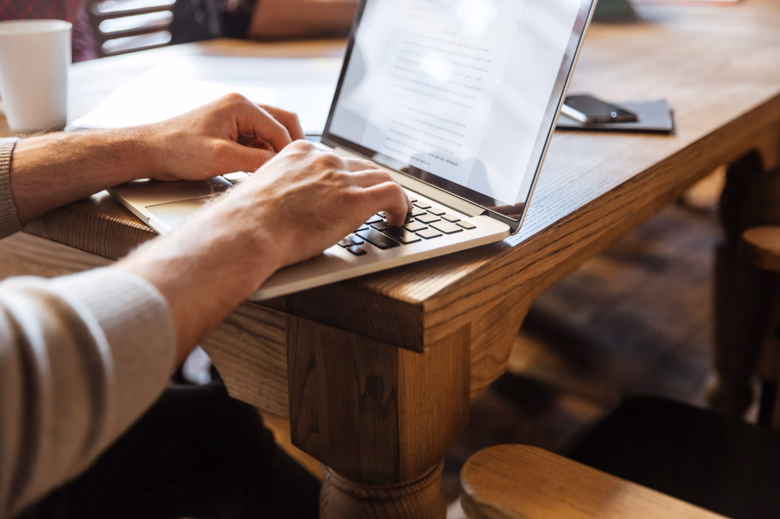Hands typing on a laptop keyboard at a wooden table with a smartphone and notebook nearby.