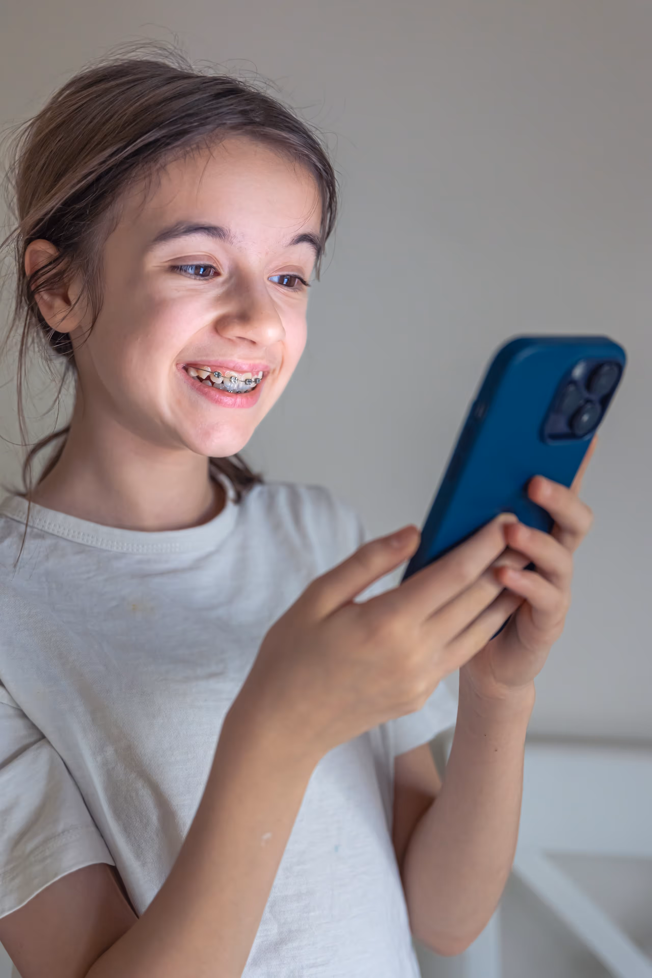 Smiling girl with braces looking at a smartphone in a light-colored room.