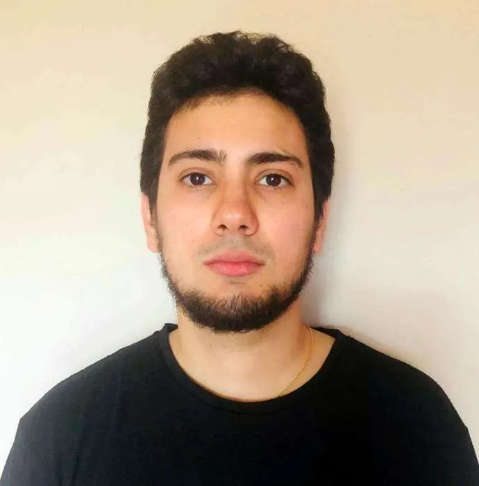 Portrait of a young man with short dark hair and a trimmed beard wearing a black shirt against a plain beige background.