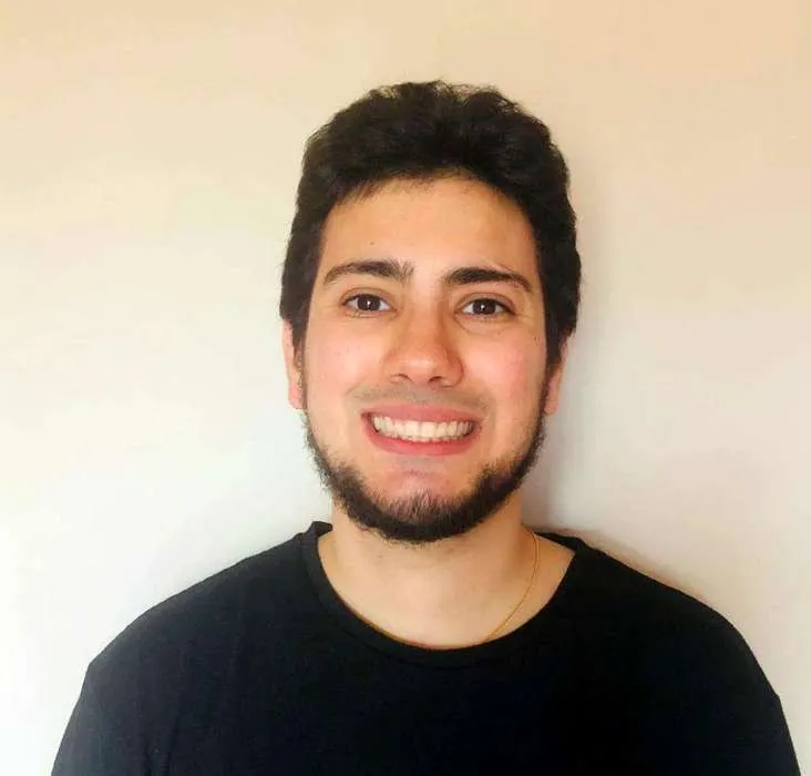 Young man with dark hair and beard smiling while wearing a black shirt against a plain light background.