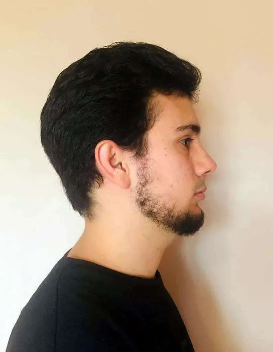 Profile of a young man with dark hair and a short beard wearing a black shirt, against a plain light background.
