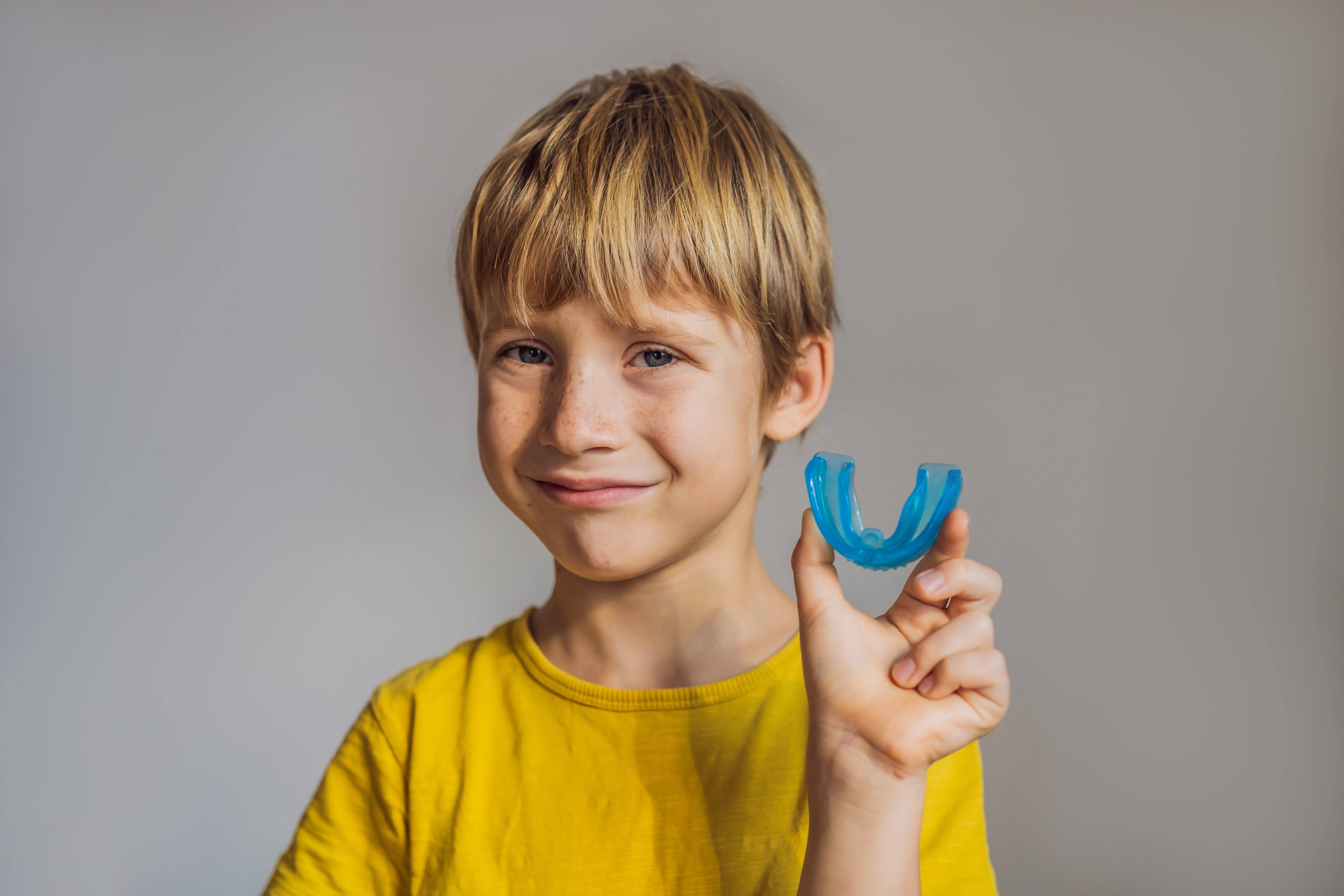 Smiling blonde boy in a yellow shirt holding a blue dental mouthguard.