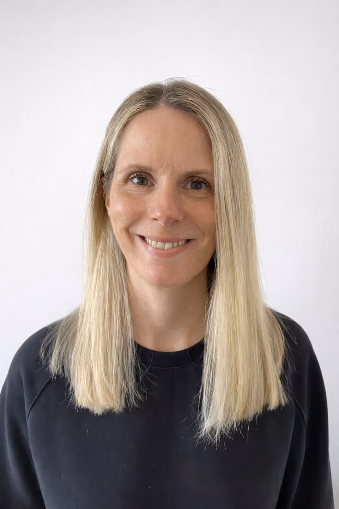 Smiling woman with long straight blonde hair wearing a black top against a plain white background.