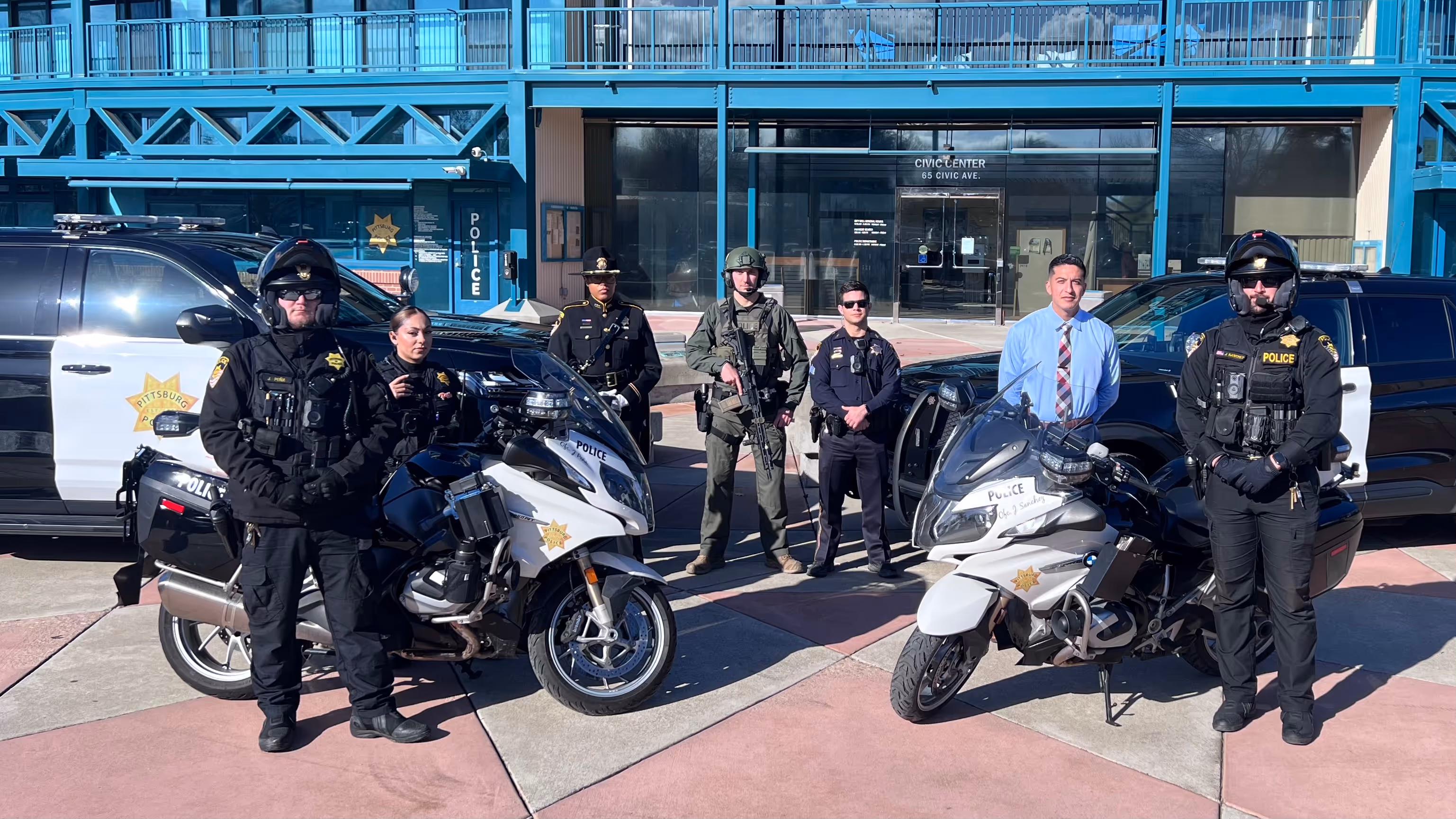 Group of police officers in uniform and tactical gear standing in front of police motorcycles and vehicles outside a civic center.