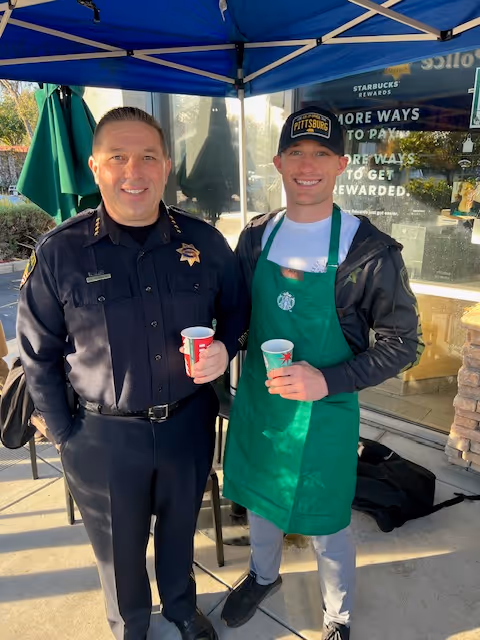 A police officer and a Starbucks barista standing side by side under a blue canopy, each holding a cup and smiling.