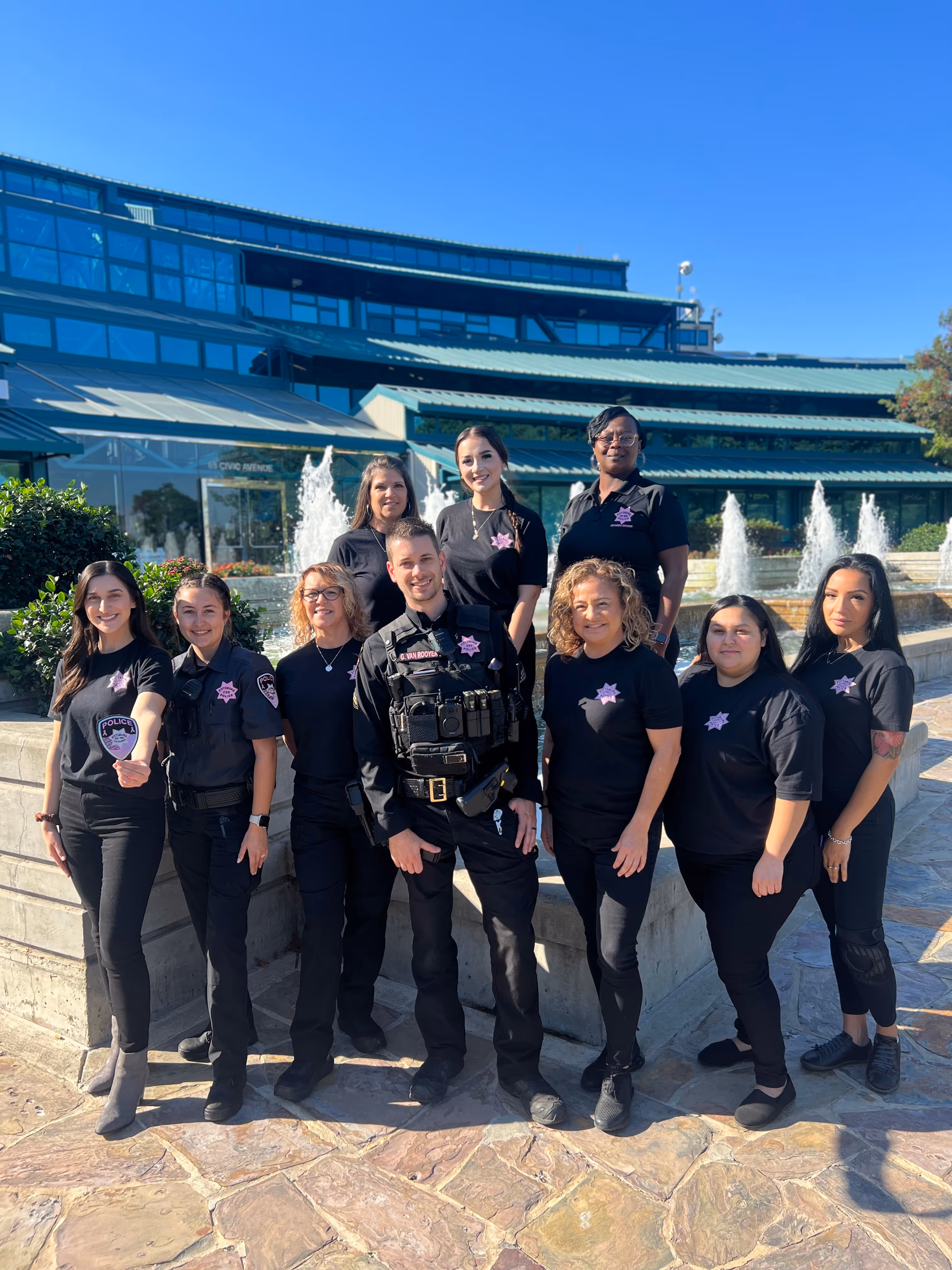 Group of ten police officers standing outside near a fountain and modern building, wearing black uniforms with pink police badges.