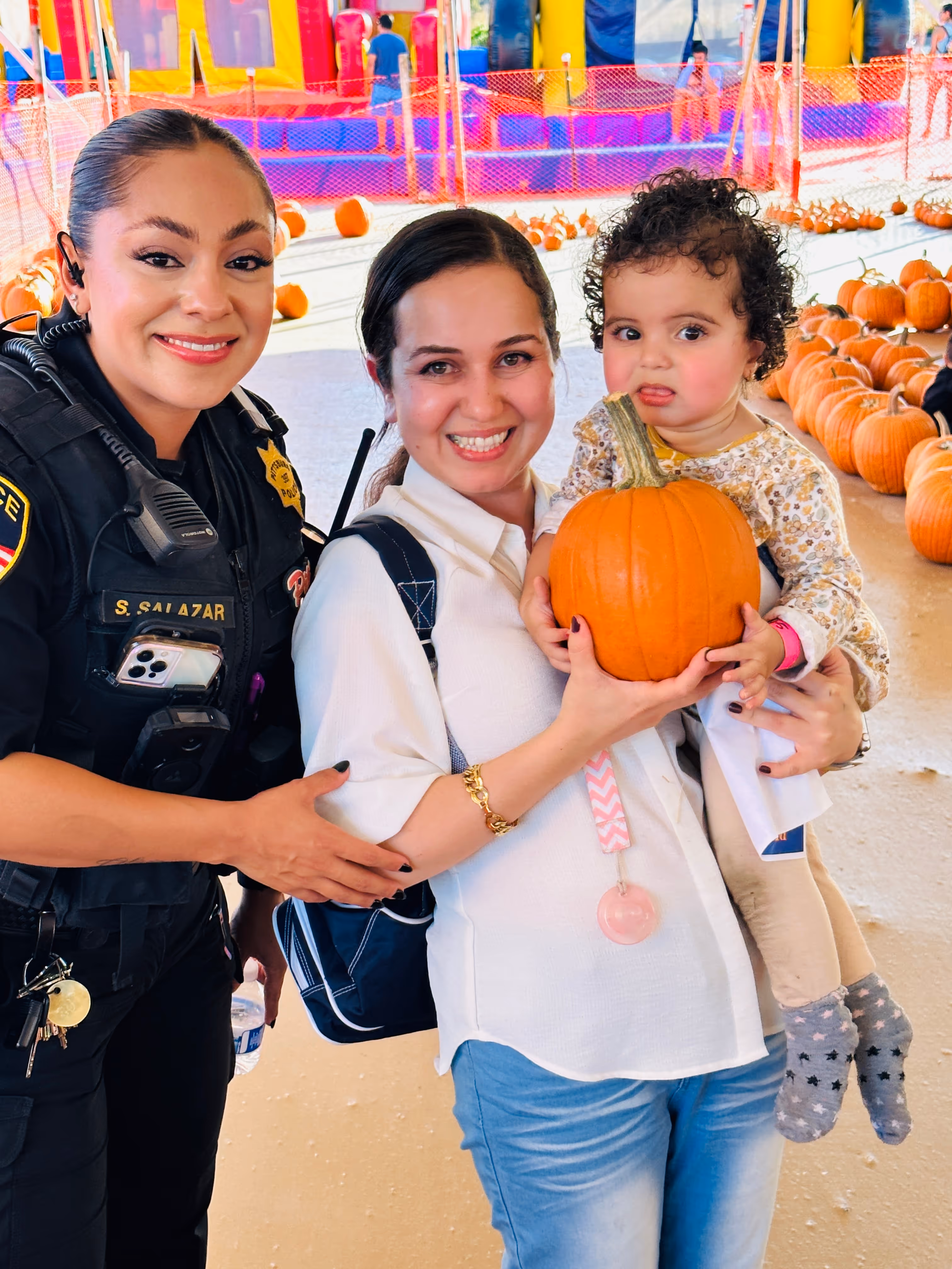 Smiling female police officer with woman holding a toddler carrying a pumpkin at a pumpkin patch.
