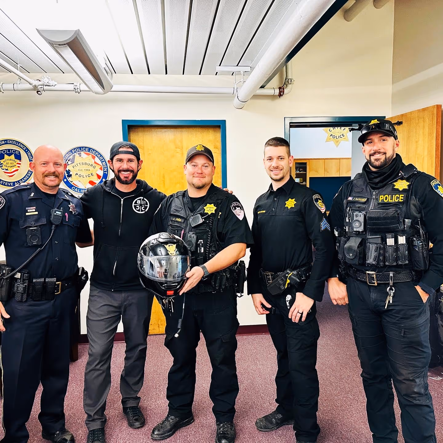 Group of five men smiling indoors, including four police officers in uniform and one casually dressed man with a backwards cap.