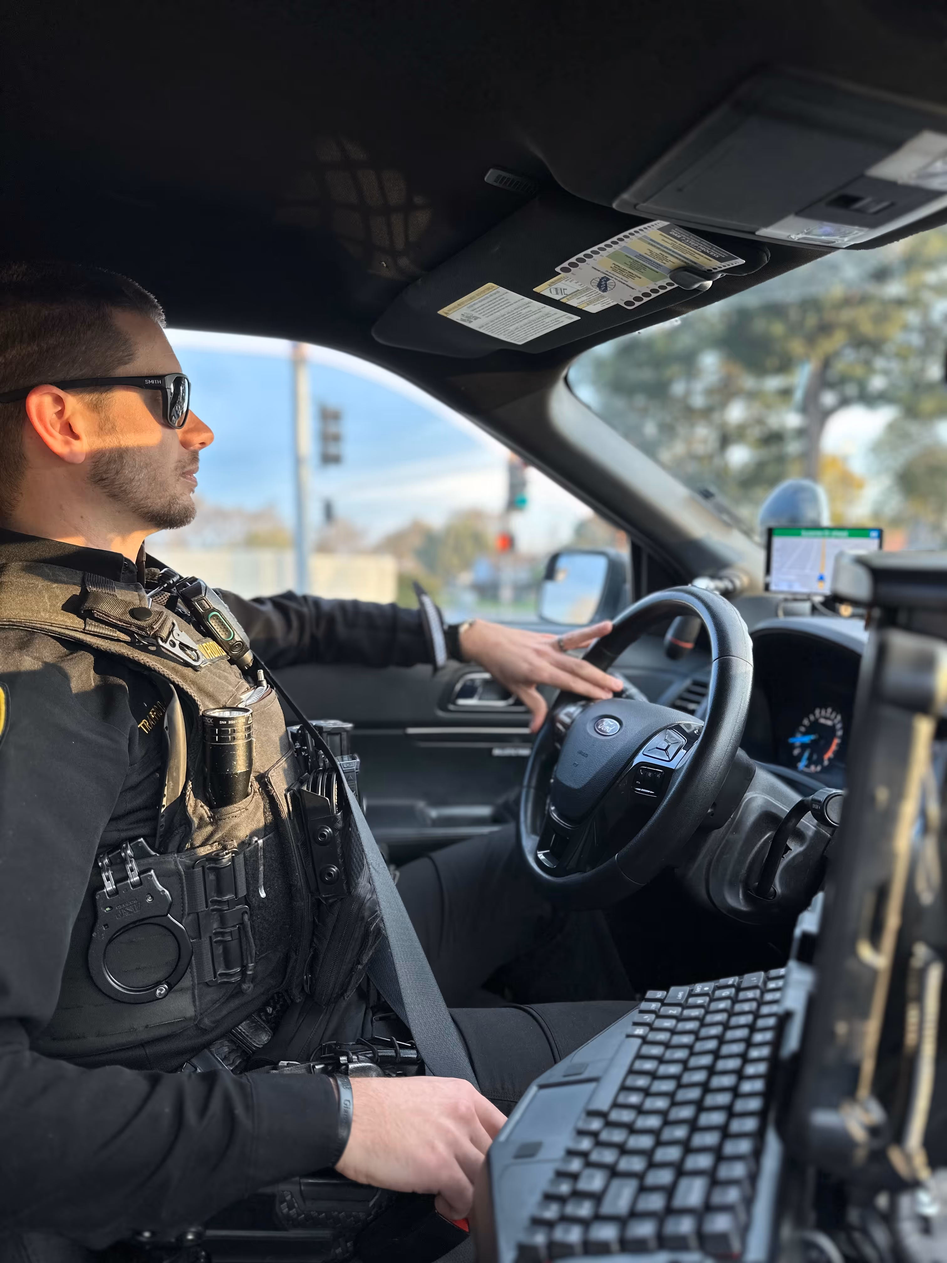 Police officer wearing sunglasses and tactical gear driving a patrol car equipped with a computer keyboard and GPS device.
