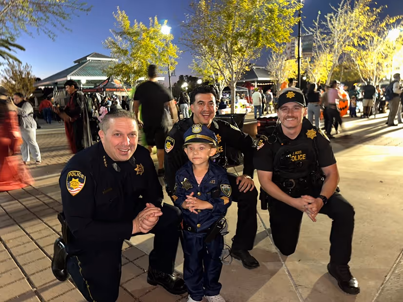 Three police officers kneeling and smiling with a young child dressed in a police uniform at an outdoor evening event.