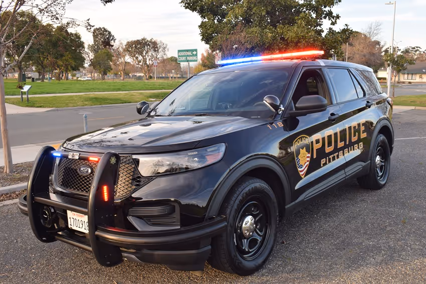 Black Pittsburg police SUV with red and blue lights flashing, parked on a street near a park.