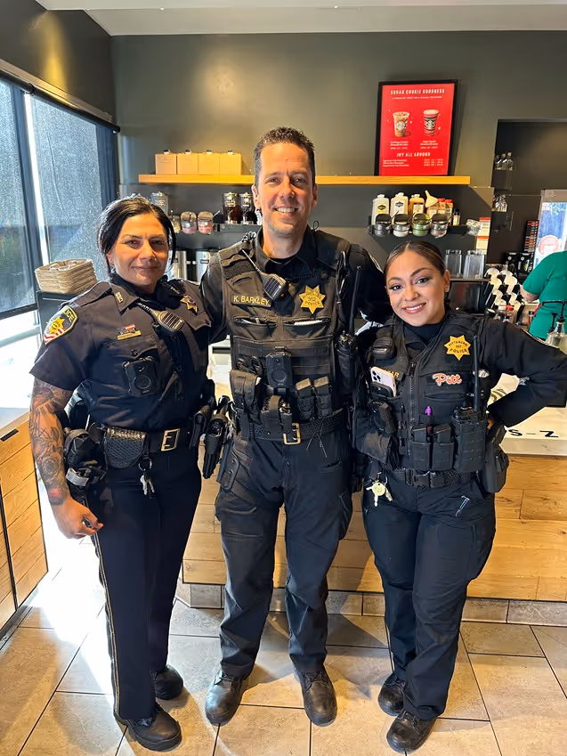 Three smiling police officers in uniform standing inside a coffee shop with wooden counters and shelves in the background.