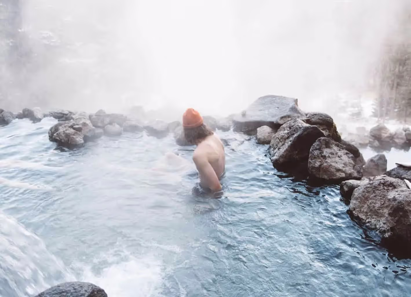 Man wearing orange winter hat sitting in steamy hot spring on mountain