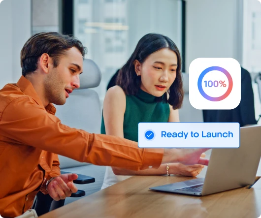 Two coworkers at desk looking at laptop with 100% progress and ‘Ready to Launch’ notification.