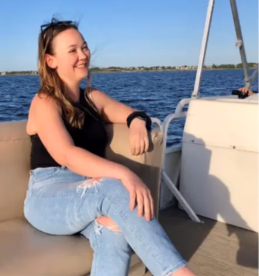 Smiling woman with sunglasses on her head sitting on a boat seat by the water, wearing a black tank top and ripped blue jeans.