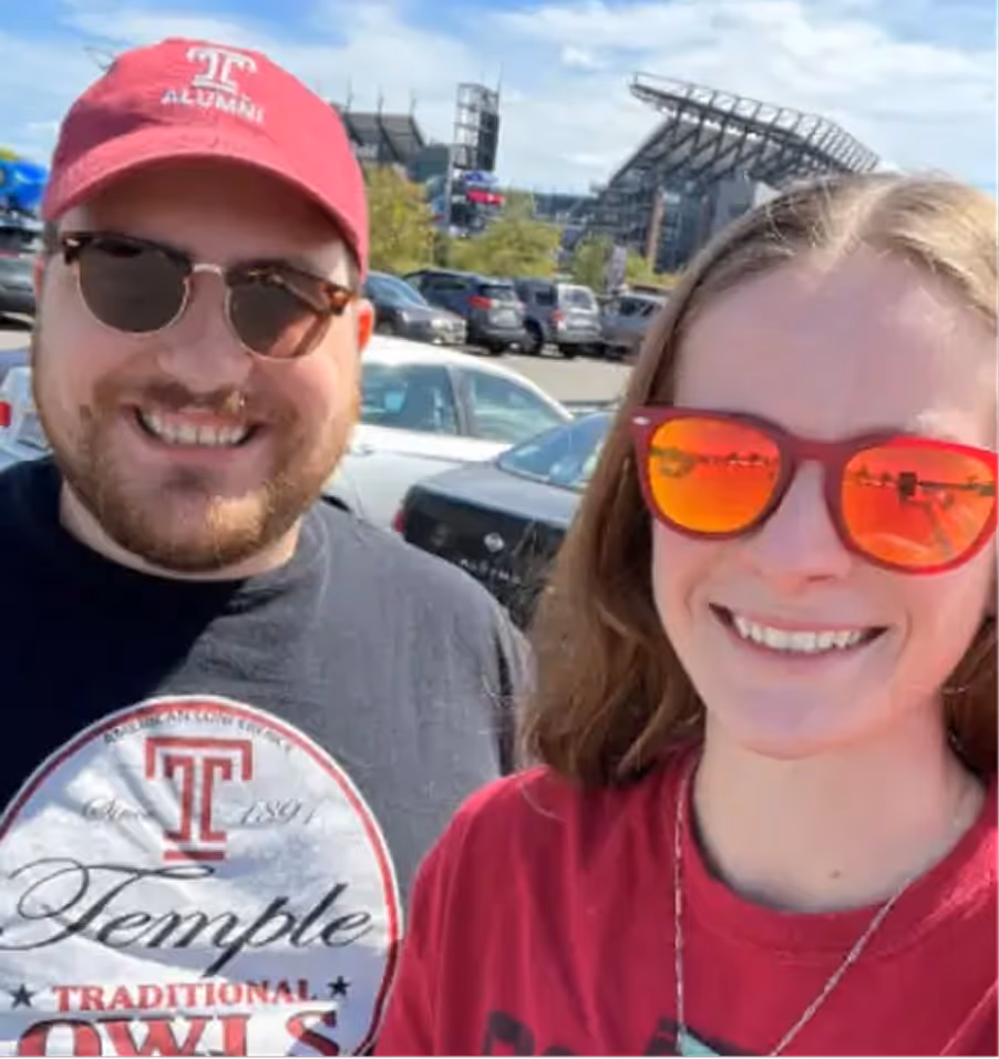 Two people smiling outdoors in a parking lot with a stadium in the background; one wearing a red Temple Alumni hat and black Temple Owls shirt, the other wearing red reflective sunglasses and a red shirt.