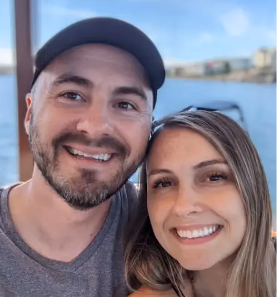 Smiling man wearing a black cap and a woman with long hair posing together outdoors near water.