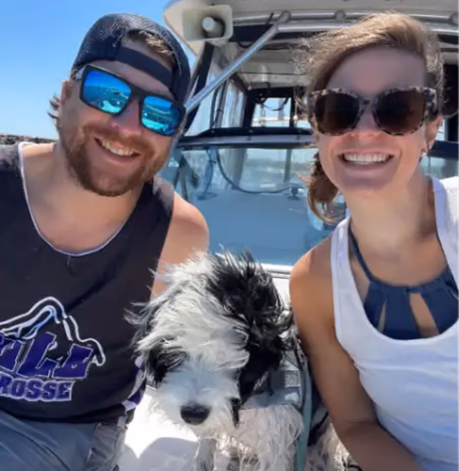 Smiling man and woman wearing sunglasses on a boat with a black and white dog between them.