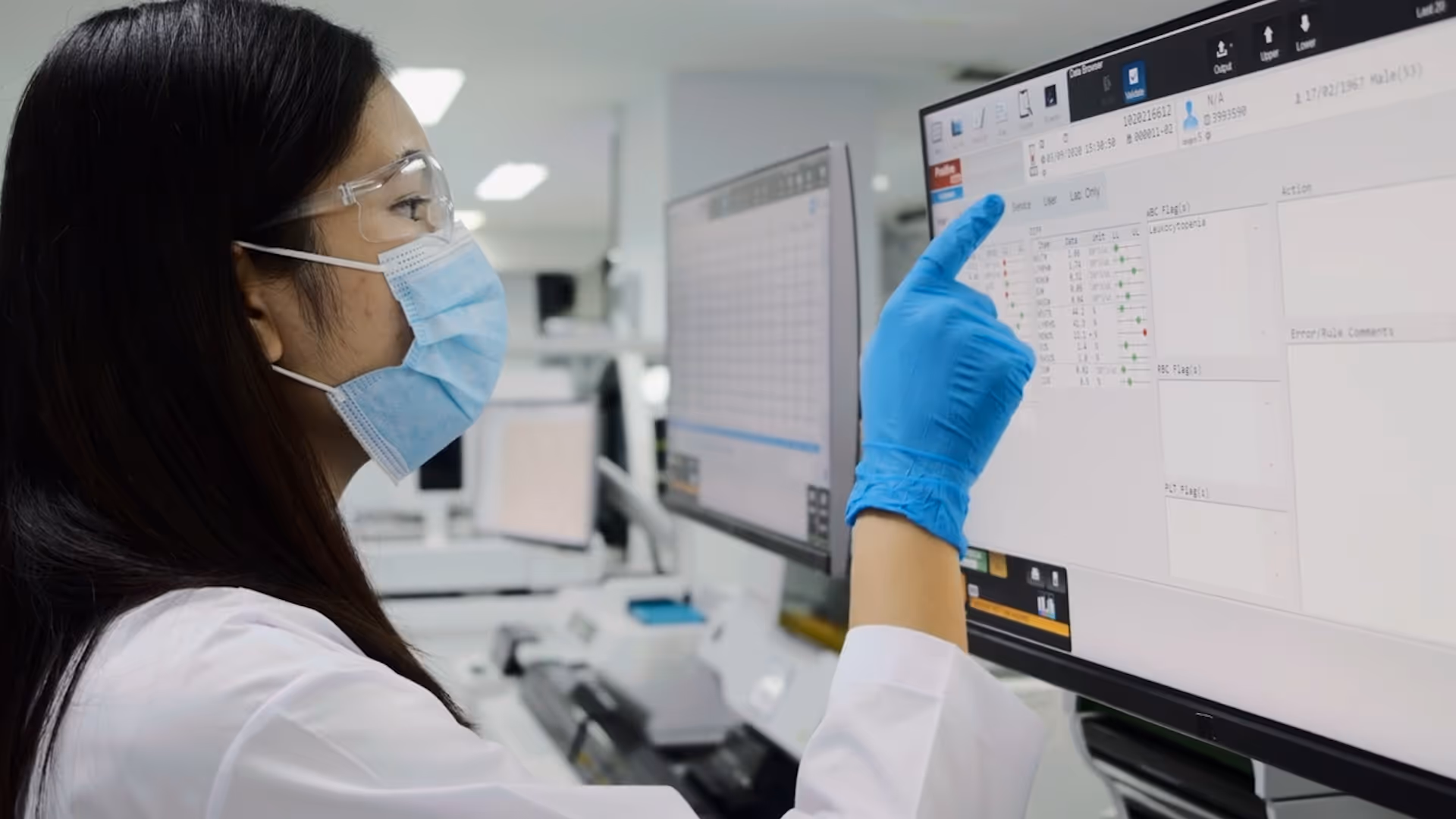 Lab technician wearing a mask, safety goggles, and blue gloves pointing at data on a computer screen in a laboratory.