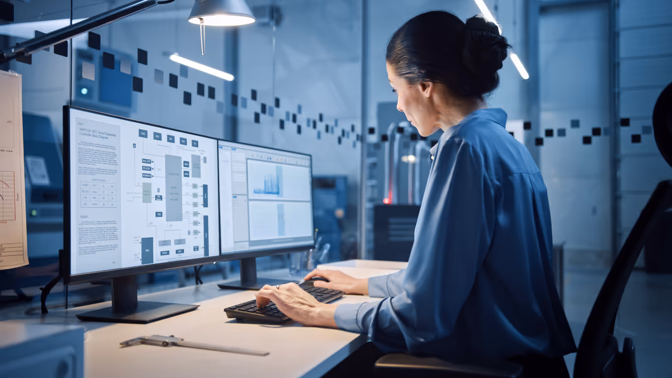 Woman in a blue shirt working on a computer with dual monitors displaying MES systems in a modern office.