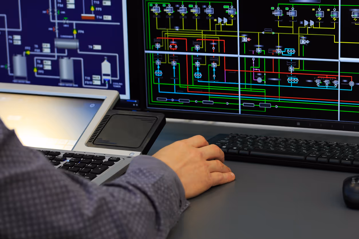 Person working at a desk with a laptop and a large monitor showing complex flow diagrams and circuit schematics.