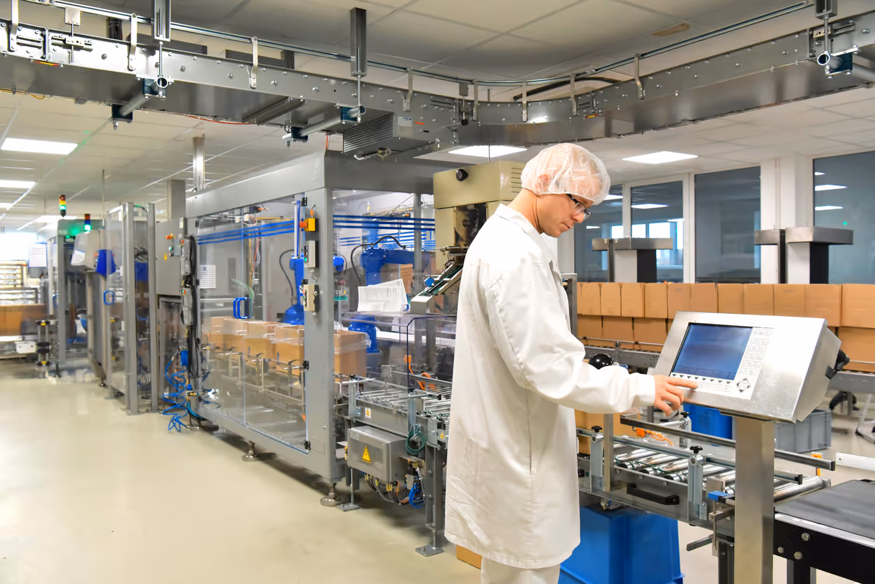 Worker in white lab coat and hairnet operating control panel in a packaging factory with conveyor belt and boxes.