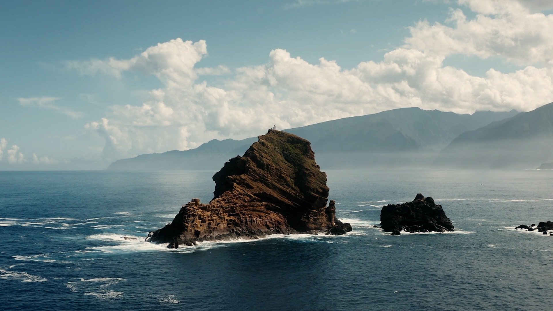 Rocky island surrounded by ocean waves with mountainous coastline and cloudy sky in the background.