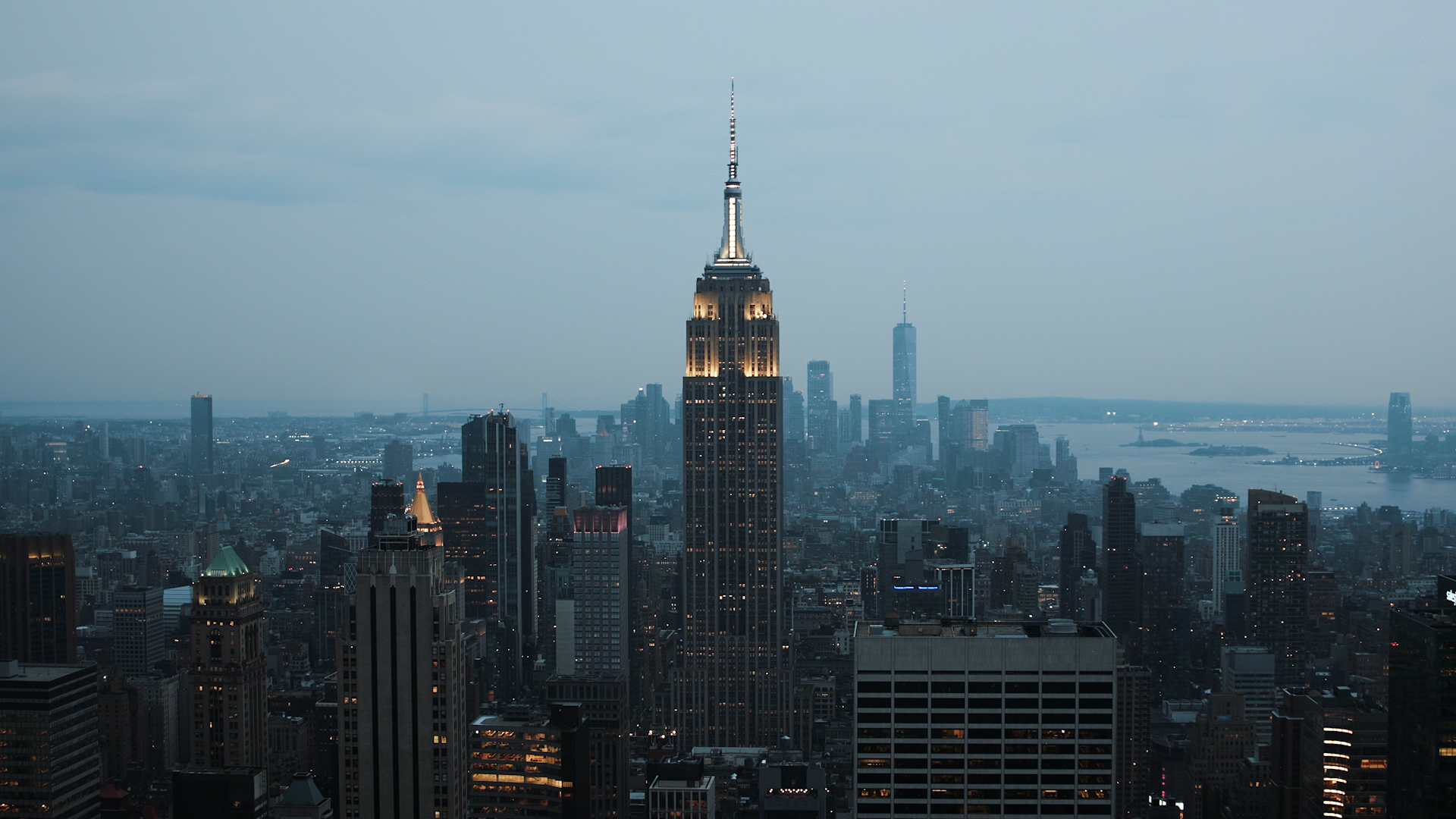 Evening skyline of New York City with the Empire State Building illuminated in the center and surrounding skyscrapers under a cloudy sky.