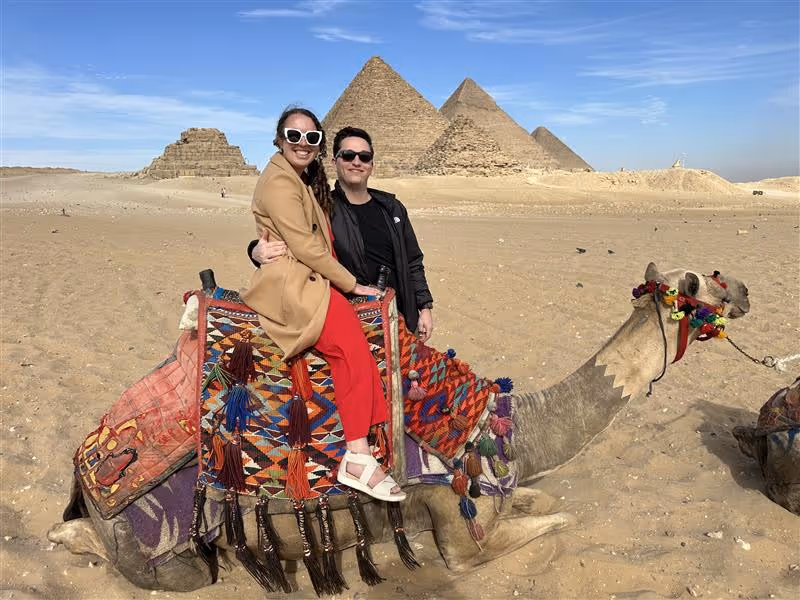 Smiling woman with long brown hair with lighter highlights wearing a dark top against a neutral background.