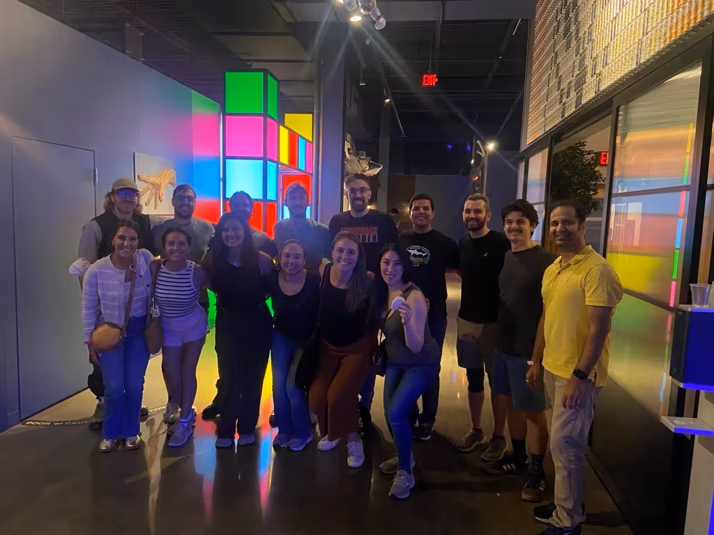 Group of fourteen people posing indoors with colorful illuminated blocks in the background.
