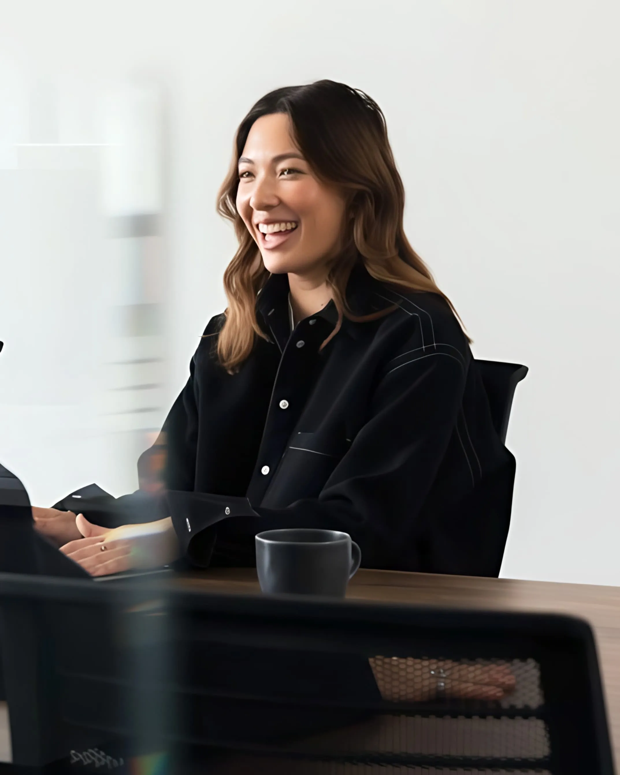 Smiling woman with wavy brown hair wearing a black shirt sitting at a table with a black mug.