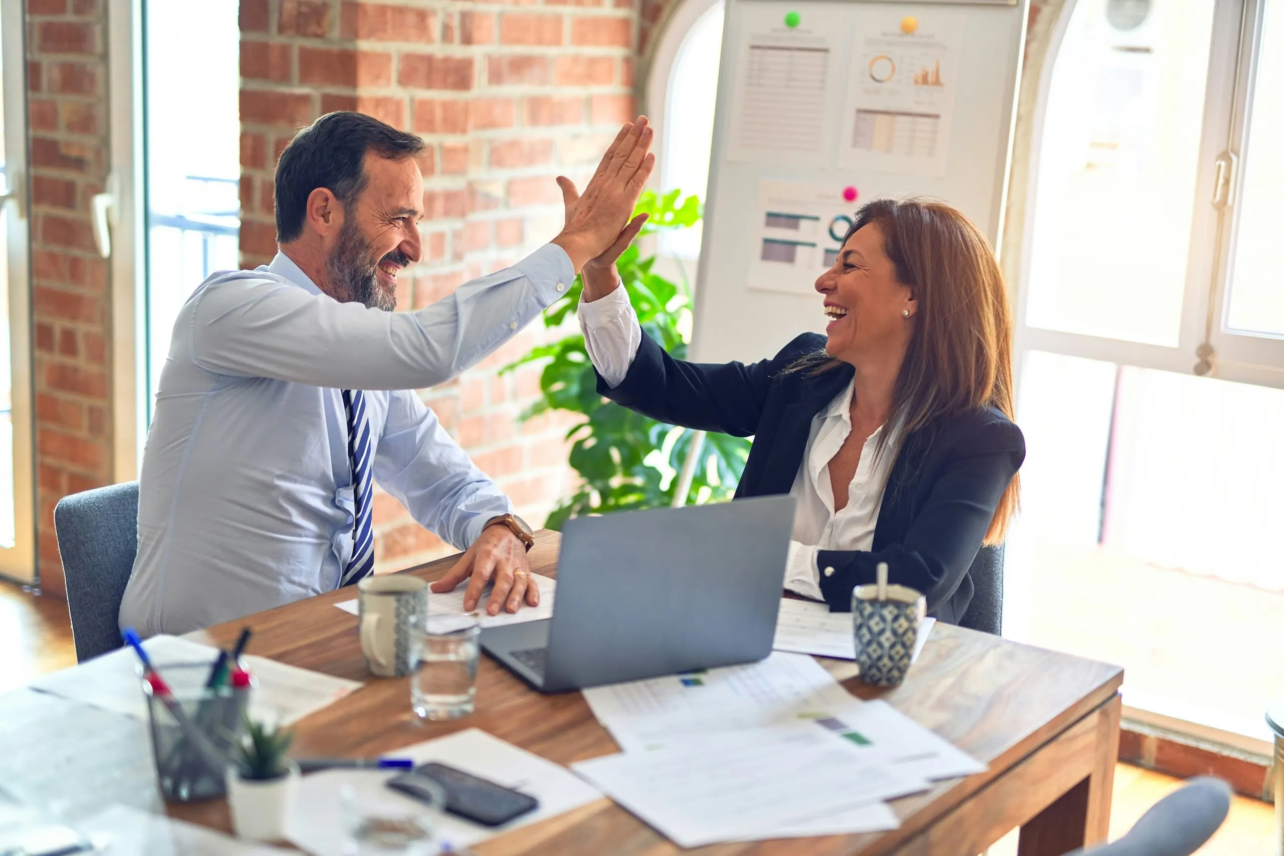 Two colleagues in business attire smiling and giving a high five over a wooden desk with paperwork and a laptop.