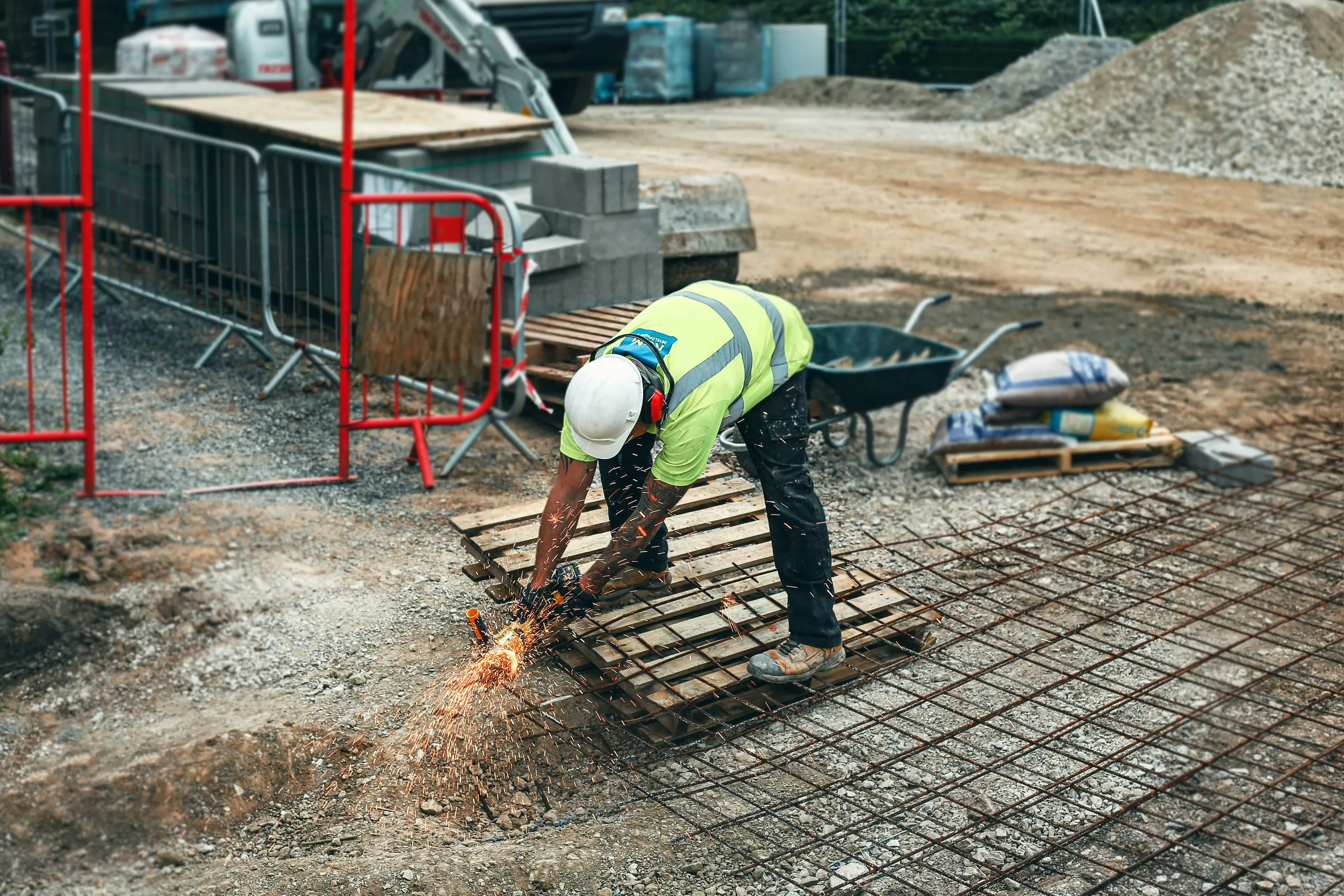 Construction worker in a hard hat and high-visibility vest cutting metal rebar with an angle grinder sparking on a construction site.