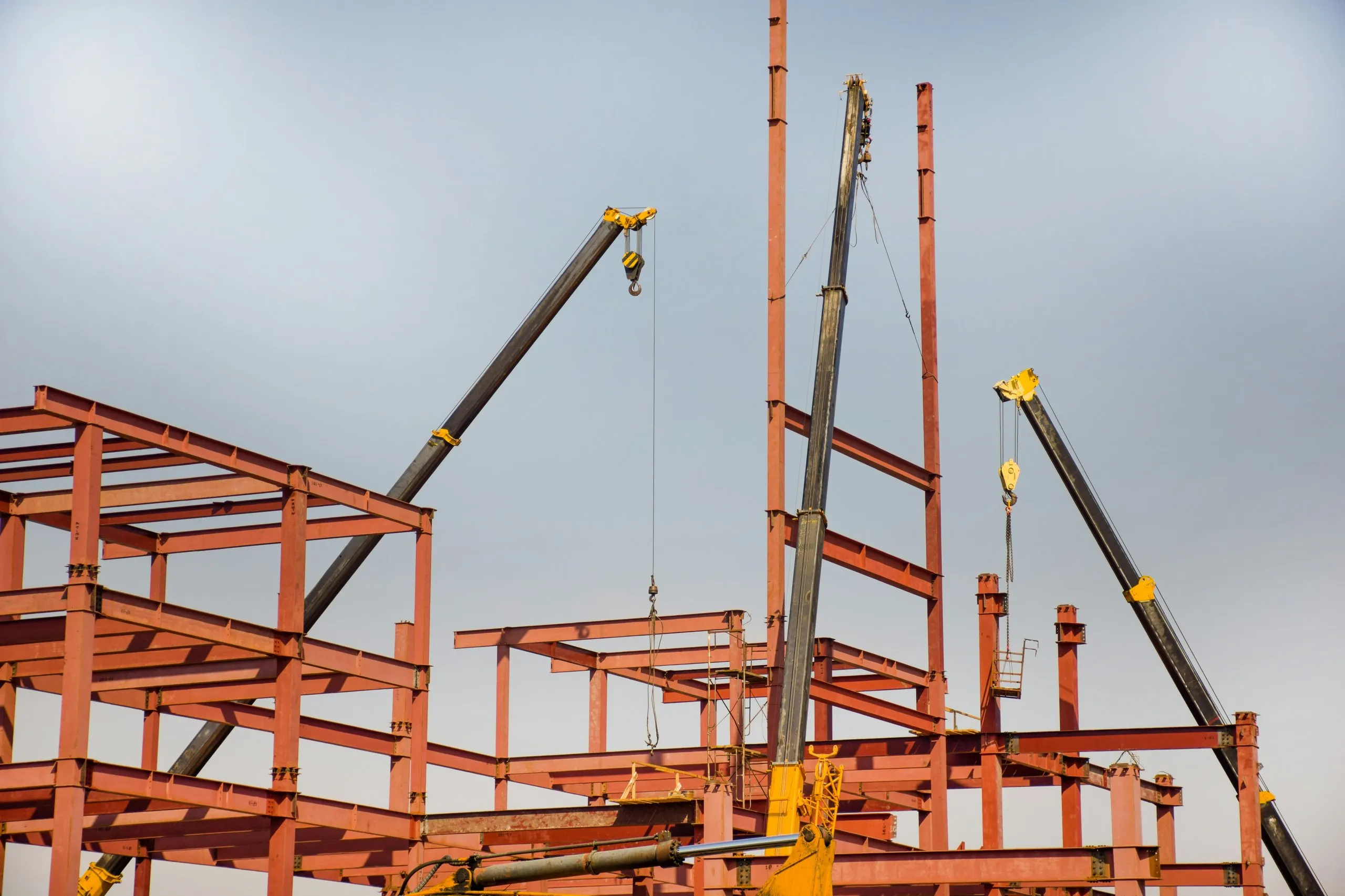 Yellow cranes lifting steel beams at a construction site with red steel framework under a clear sky.