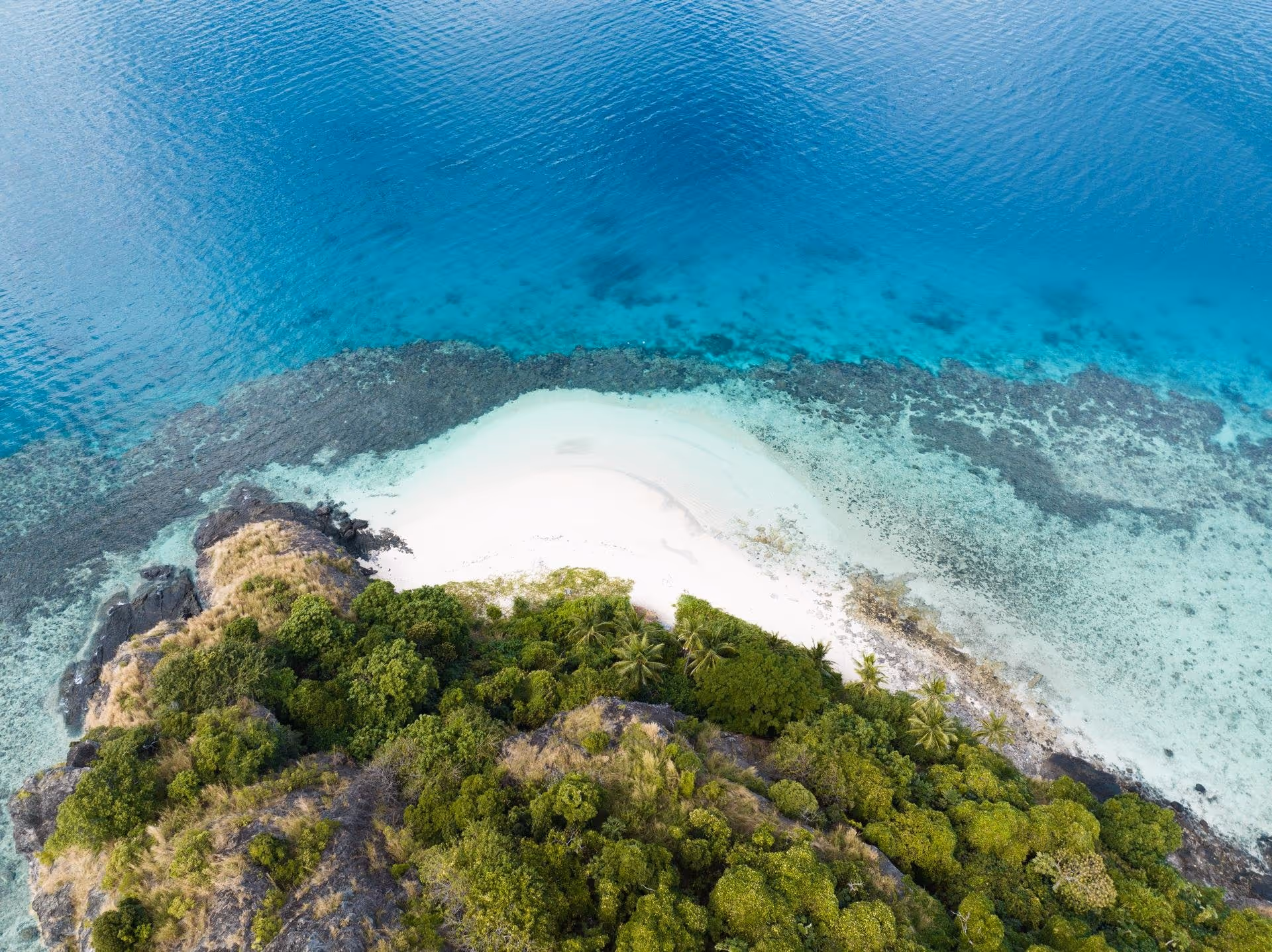 Aerial view of a small tropical island with dense green vegetation, white sandy beach, and clear turquoise ocean water.