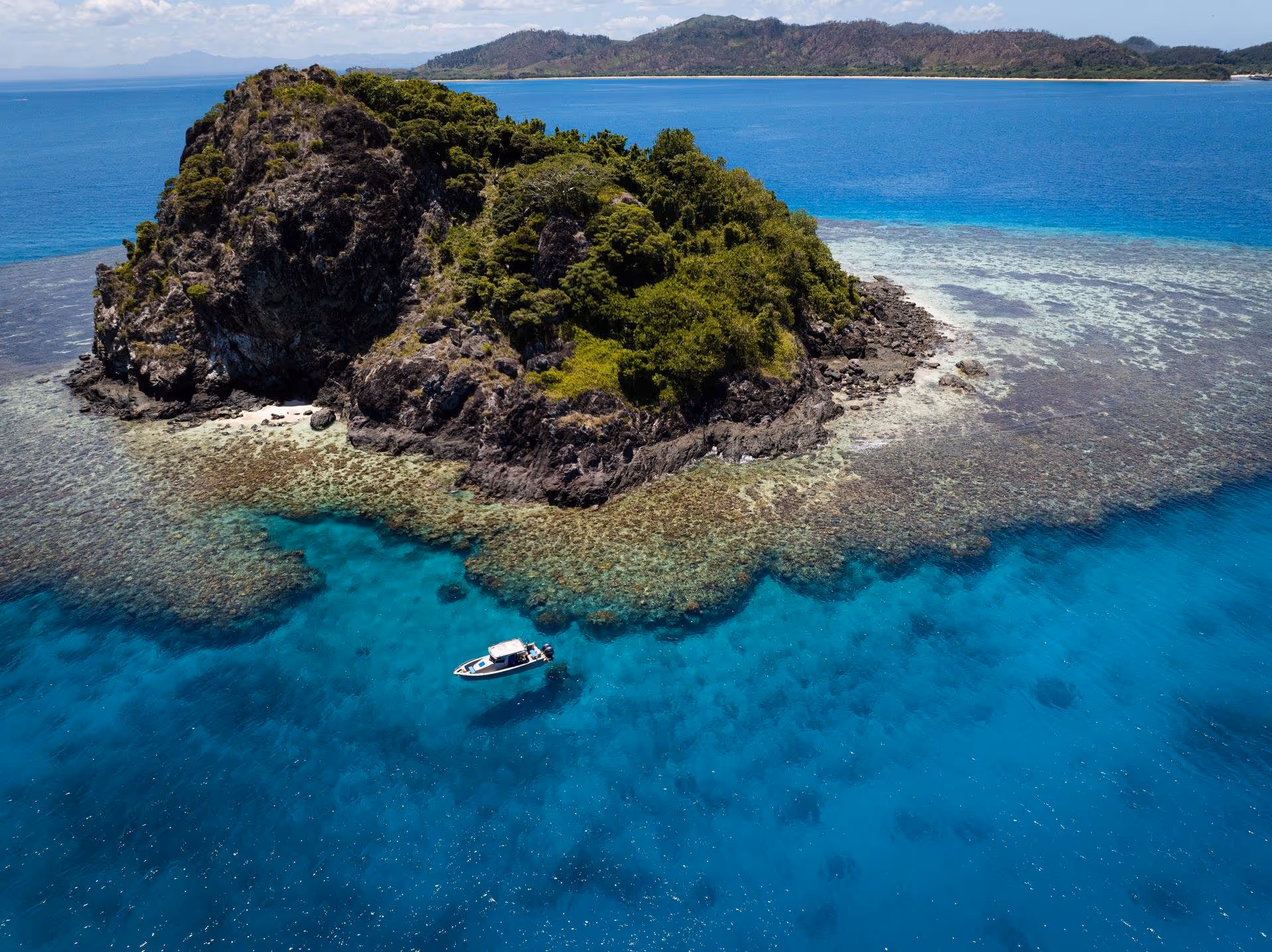 Small rocky island covered with green vegetation surrounded by clear blue ocean with a boat floating nearby.