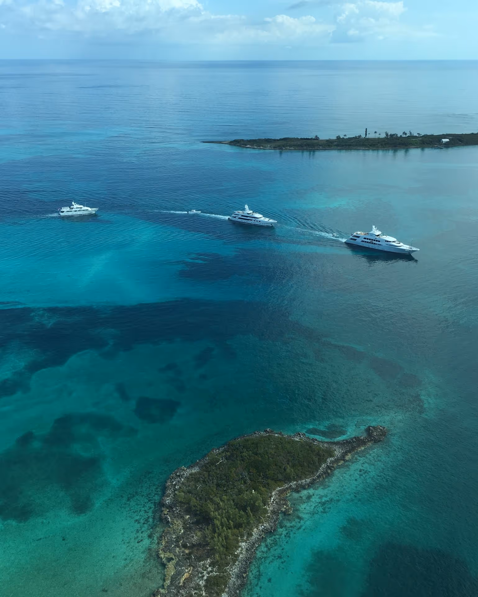 Aerial view of three white yachts sailing in clear turquoise waters near small green islands under a partly cloudy sky.