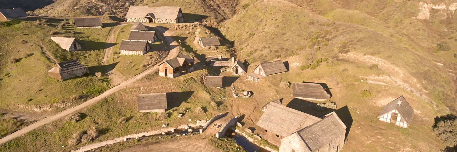 Aerial view of a small military outpost with sandbag walls on a grassy hill surrounded by rolling green mountains under a partly cloudy sky.