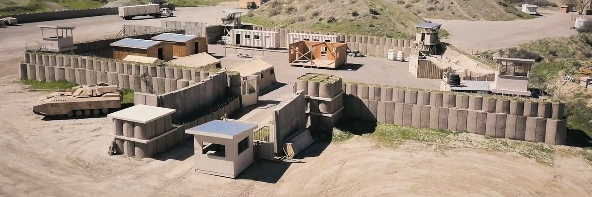 Aerial view of a small military outpost with sandbag walls on a grassy hill surrounded by rolling green mountains under a partly cloudy sky.