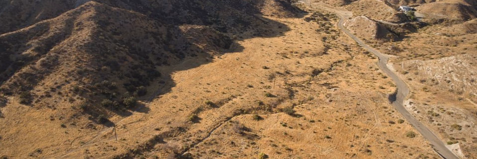 Aerial view of a small military outpost with sandbag walls on a grassy hill surrounded by rolling green mountains under a partly cloudy sky.