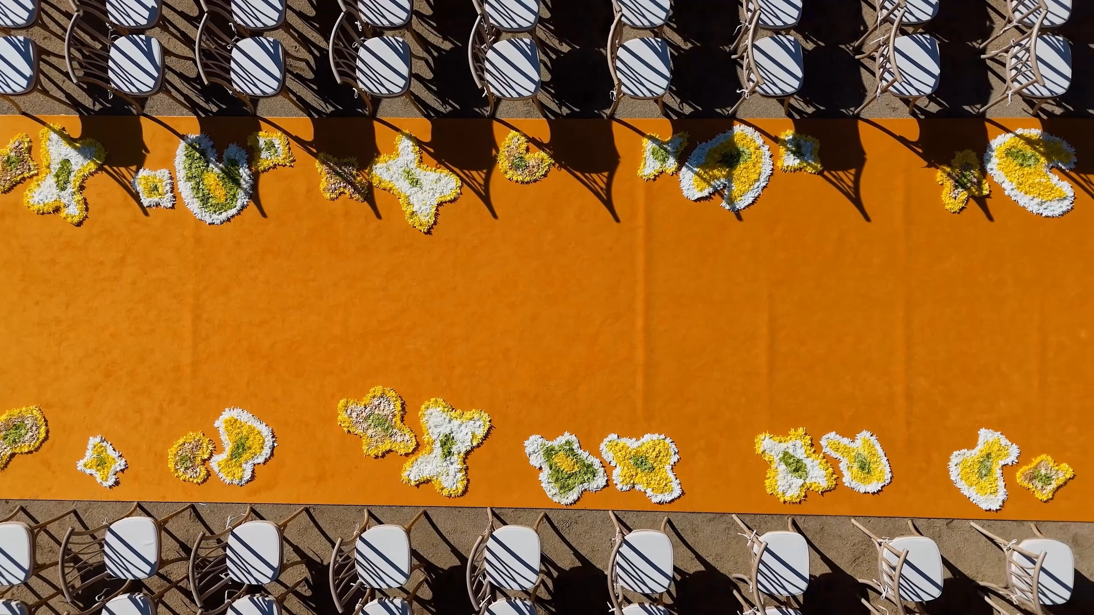 Overhead view of an orange aisle runner with white and yellow floral arrangements on both edges, and rows of chairs casting striped shadows.