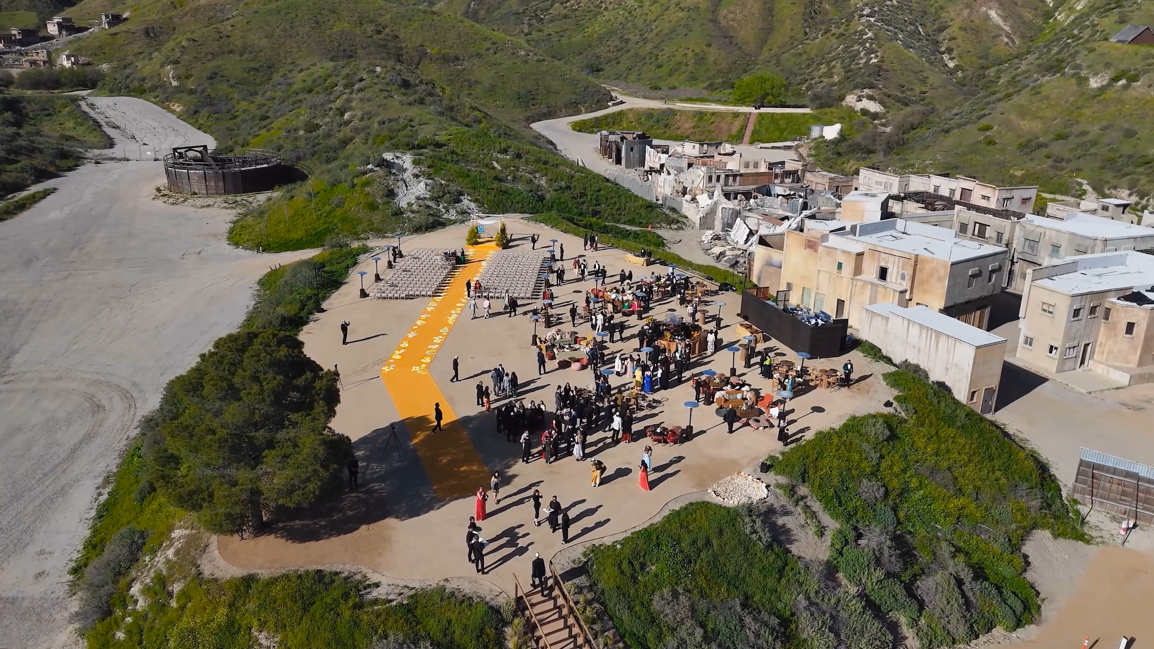 Aerial view of an outdoor event setup with chairs, tables, and guests on a concrete platform surrounded by green hills and old buildings.