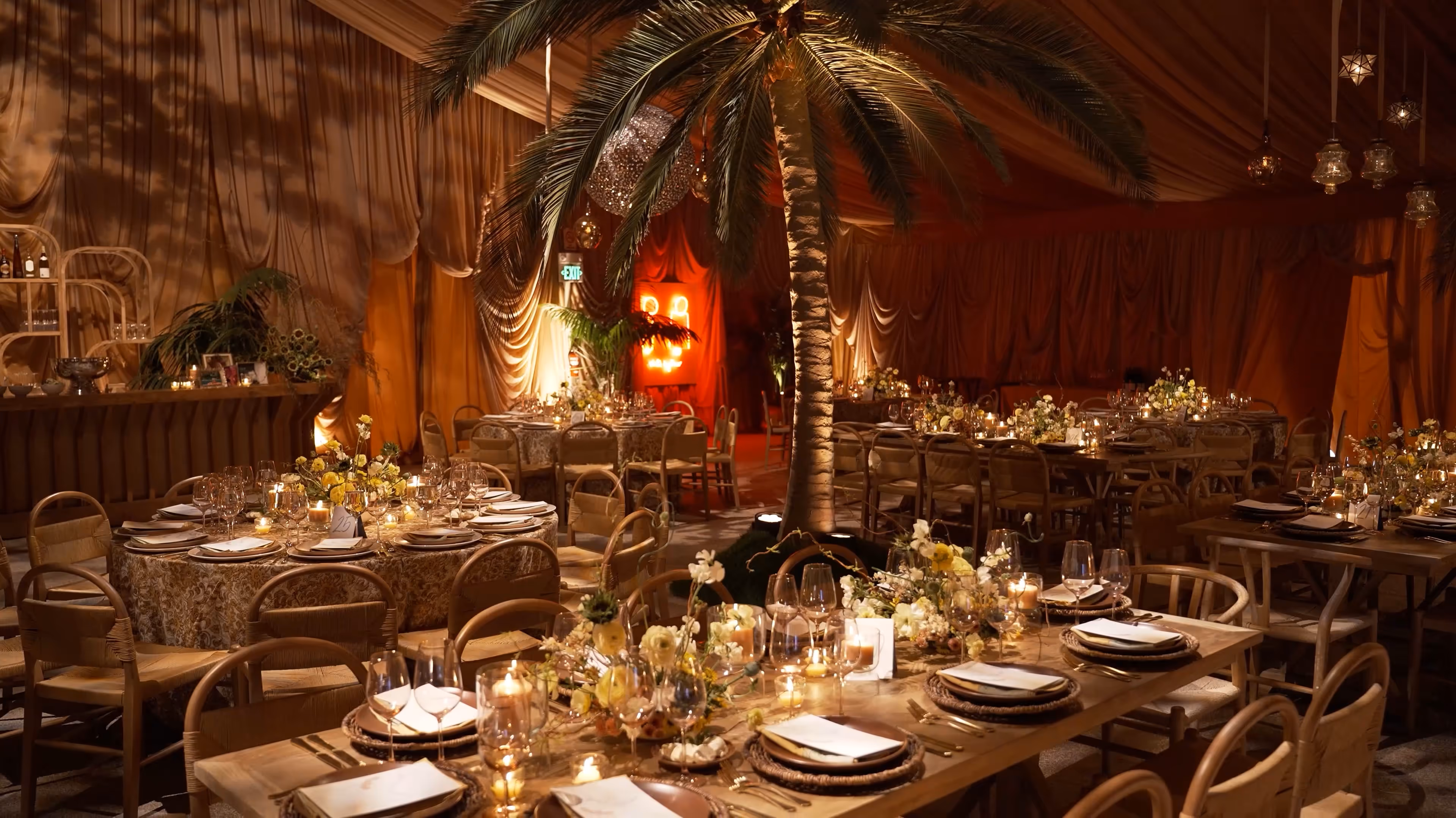 Elegant indoor event space with round and rectangular tables set with plates, glasses, floral centerpieces, and candles under draped ceilings and a tall palm tree.